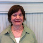 Nancy Brumback - white woman with short brown hair, wearing a green collared shirt, standing in front of a white wall