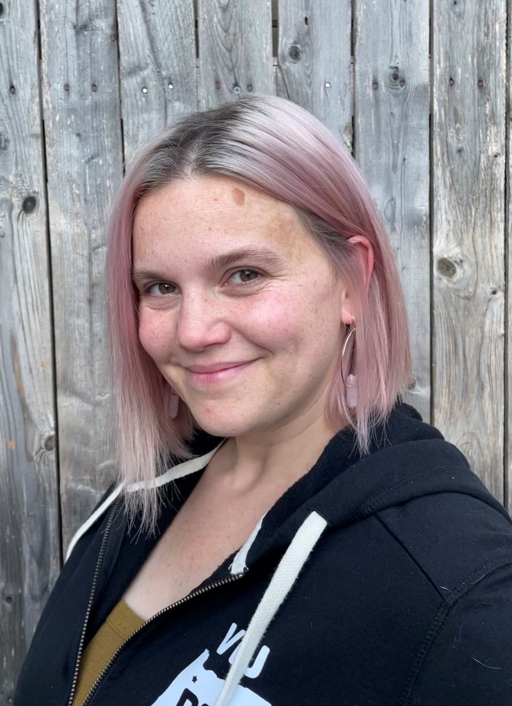 Amelia Fowler - white woman with short pink hair, wearing a black hoodie, standing in front of a wooden fence.