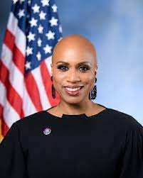 Congresswoman Ayanna Pressley - Black woman with no hair, wearing a black shirt, sitting in front of an American flag