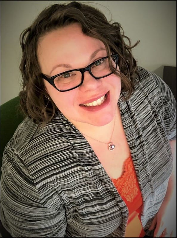 Michelle Bishop - white woman with short curly brown hair, wearing black rimmed glasses and a black and white stripped sweater over an orange shirt.