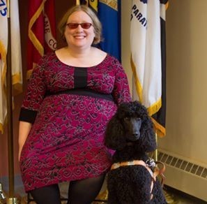 Rachel Tanenhaus - white woman with short hair, wearing black rimmed glasses and a red and black floral dress, sitting next to a black poodle and in front of multiple flags.