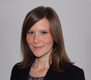 Tatum Pritchard - white woman with medium length brown hair, wearing a black blazer and standing in front of a white wall